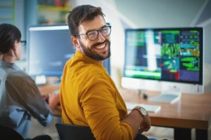 Closeup over the shoulder view of a cheerful mid 20's software developer at the office smiling to the camera. His female coworker is next him, still focused and working.